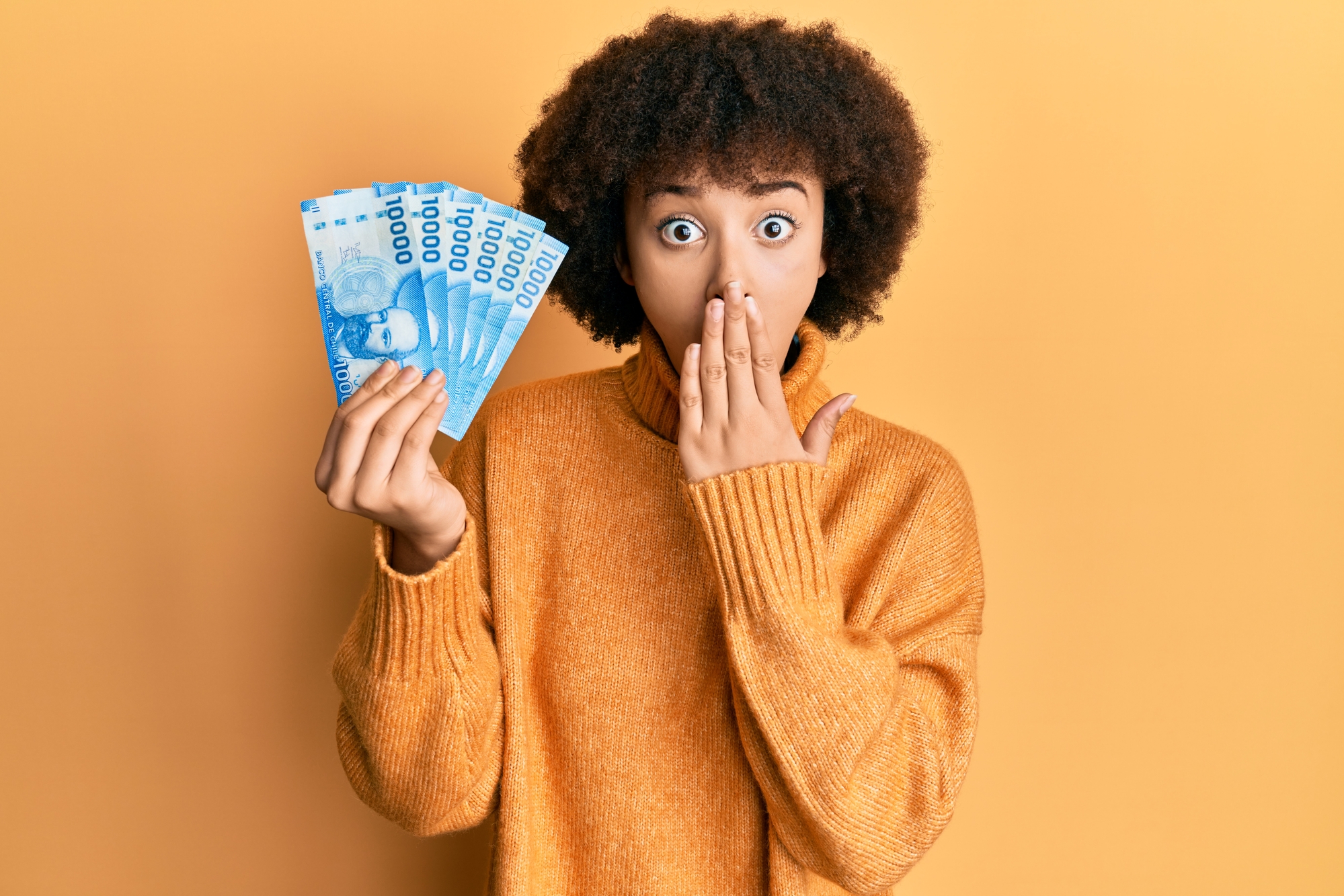 Young hispanic girl holding 10000 chilean pesos covering mouth with hand, shocked and afraid for mistake. surprised expression